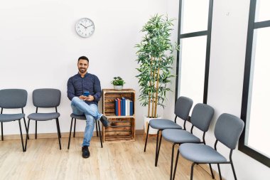 Young hispanic man using smartphone sitting on chair at waiting room
