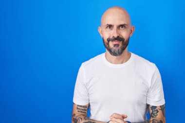 Mavi arka planda elleri birarada duran ve gülümseyen gevşemiş ve neşeli İspanyol dövmeli bir adam. Başarı ve iyimserlik 