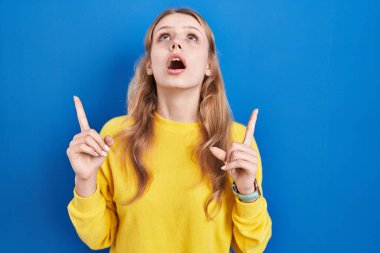 Mavi arka plan üzerinde duran genç beyaz kadın şaşırmış ve şaşırmış bir şekilde yukarı bakıyor ve parmaklarıyla işaret ediyor ve kollarını kaldırıyor.. 
