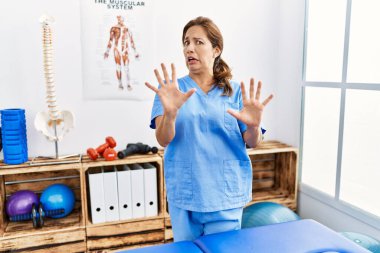 Middle age hispanic physiotherapist woman working at pain recovery clinic afraid and terrified with fear expression stop gesture with hands, shouting in shock. panic concept. 