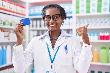 African woman with dreadlocks working at pharmacy drugstore holding credit card pointing thumb up to the side smiling happy with open mouth 