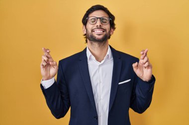 Handsome latin man standing over yellow background gesturing finger crossed smiling with hope and eyes closed. luck and superstitious concept. 