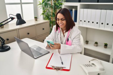 Young beautiful hispanic woman doctor using smartphone working at clinic