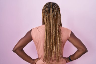 Saçları örülmüş, pembe arka planda dikilmiş, kolları vücutta geriye doğru bakan Afro-Amerikalı kadın. 
