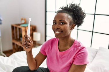 Elektrikli diş fırçası tutan Afrikalı Amerikalı kadın yatak odasında oturuyor.