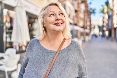 Orta yaşlı sarışın kadın sokakta kendinden emin bir şekilde gülümsüyor.