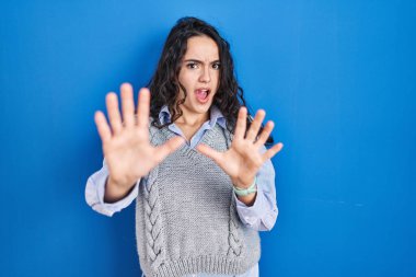 Mavi arka planda duran esmer kadın korku ifadesinden korkmuş ve dehşete düşmüş. El hareketlerini durdur, şok içinde bağır. panik konsepti. 