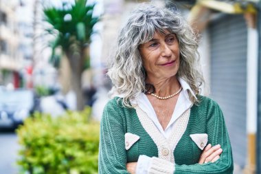 Orta yaşlı kadın gülümsüyor. Kollarını kavuşturup caddede duruyor.