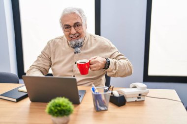 Middle age grey-haired man business worker using laptop drinking coffee at office