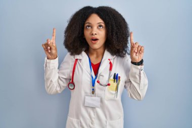 Genç Afro-Amerikan kadın doktor üniforması ve steteskop giyiyor. Şaşırmış ve şaşırmış bir şekilde yukarı bakıyor ve parmaklarıyla işaret ediyor ve kollarını kaldırıyor.. 