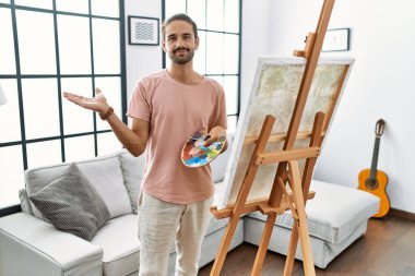 Young hispanic man with beard painting on canvas at home pointing aside with hands open palms showing copy space, presenting advertisement smiling excited happy 