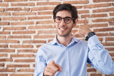 Young hispanic man standing over brick wall background smiling doing talking on the telephone gesture and pointing to you. call me. 
