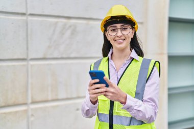 Young hispanic woman architect using smartphone at street