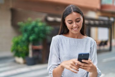 Genç, güzel, İspanyol bir kadın sokakta akıllı telefon kullanarak gülümsüyor.