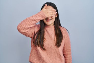 Young latin woman standing over blue background smiling and laughing with hand on face covering eyes for surprise. blind concept. 