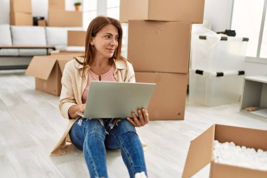 Orta yaşlı, beyaz bir kadın yeni evinde dizüstü bilgisayar kullanıyor..