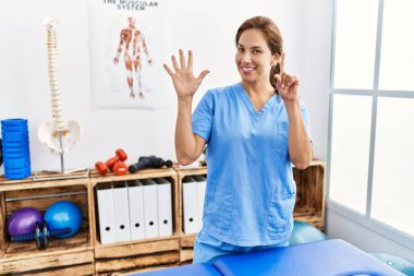 Middle age hispanic physiotherapist woman working at pain recovery clinic showing and pointing up with fingers number six while smiling confident and happy. 