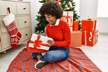 Genç Afro-Amerikan kadın boks hediyesini açıyor evde Noel ağacının yanında oturuyor.
