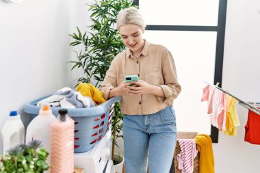 Çamaşırhanede akıllı telefon kullanan, kendine güvenen beyaz bir kadın.