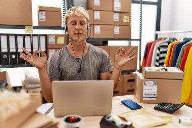 Young blond man wearing operator headset working at online shop relax and smiling with eyes closed doing meditation gesture with fingers. yoga concept. 