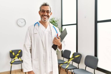 Orta yaşlı, gri saçlı, gülümseyen bir doktor. Klinik bekleme odasında kendine güvenen bir doktor.