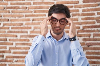 Young hispanic man standing over brick wall background with hand on head, headache because stress. suffering migraine. 