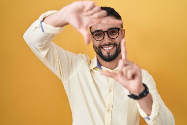 İş kıyafetleri ve gözlük takan İspanyol genç adam gülümsüyor ve mutlu yüzlü eller ve parmaklarla çerçeve yapıyor. yaratıcılık ve fotoğrafçılık kavramı. 