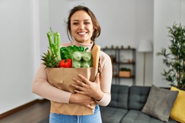 Genç, güzel, İspanyol bir kadın gülümsüyor. Elinde alışveriş torbası var.