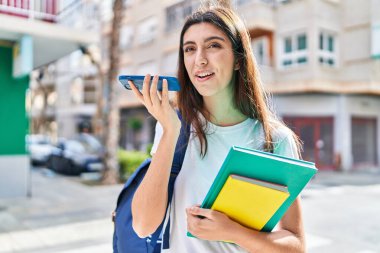 Young beautiful hispanic woman student holding books using smartphone at street