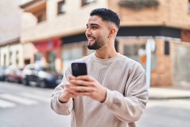 Genç Arap adam sokakta akıllı telefon kullanarak kendinden emin gülümsüyor.