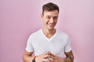 Caucasian man standing over pink background hands together and fingers crossed smiling relaxed and cheerful. success and optimistic 