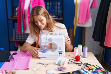 Young blonde girl tailor smiling confident using sewing machine at sewing studio