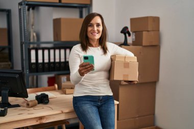 Middle age woman ecommerce business worker using smartphone holding packages at office
