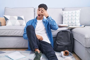 Young hispanic man sitting on the floor studying for university peeking in shock covering face and eyes with hand, looking through fingers with embarrassed expression. 
