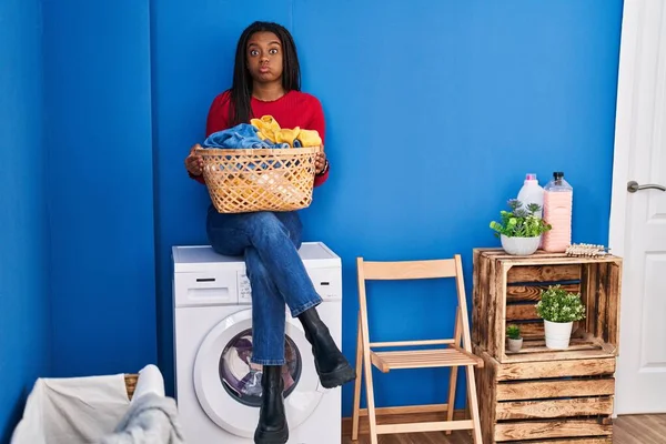 年轻的非洲裔美国人 头戴辫子 手里拿着洗衣篮 坐在洗衣机上 满脸滑稽地吹着腮帮子 嘴被空气吹胀 — 图库照片
