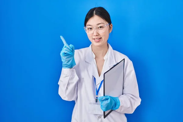 Chinese young woman working at scientist laboratory pointing with hand finger to the side showing advertisement, serious and calm face