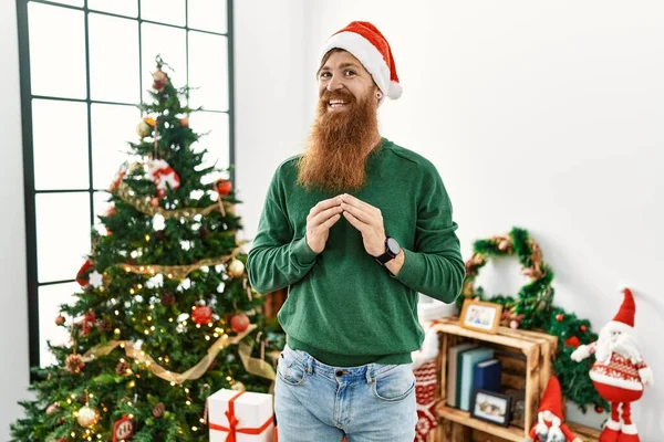 红头发的男人留着长长的胡子 手挽着圣诞树头戴圣诞帽 手指交叉在一起 微笑着 轻松而快乐 成功与乐观 — 图库照片