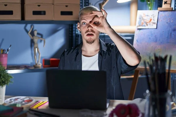 Young caucasian man using laptop at night at art studio doing ok gesture shocked with surprised face, eye looking through fingers. unbelieving expression.