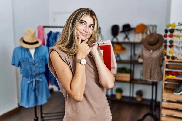 年轻的金发女人拿着购物袋在零售店里 看着相机 满心自信地微笑着 双臂交叉 双手托着下巴 积极地思考 — 图库照片
