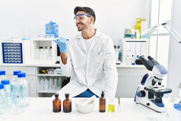 Young hispanic man working at scientist laboratory pointing thumb up to the side smiling happy with open mouth