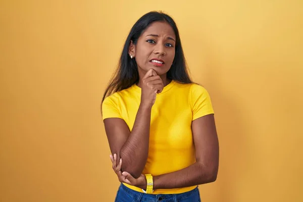 Young indian woman standing over yellow background thinking worried about a question, concerned and nervous with hand on chin