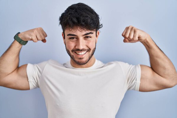 Hispanic man with beard standing over white background showing arms muscles smiling proud. fitness concept. 