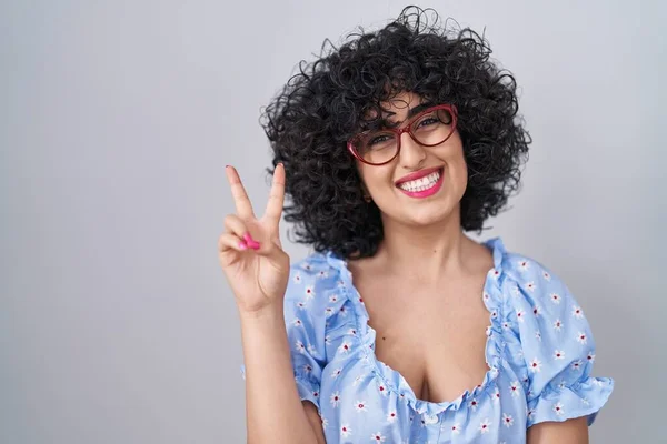 Young brunette woman with curly hair wearing glasses over isolated background smiling with happy face winking at the camera doing victory sign. number two.