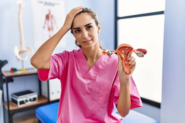 Young hispanic woman holding model of female genital organ at rehabilitation clinic stressed and frustrated with hand on head, surprised and angry face