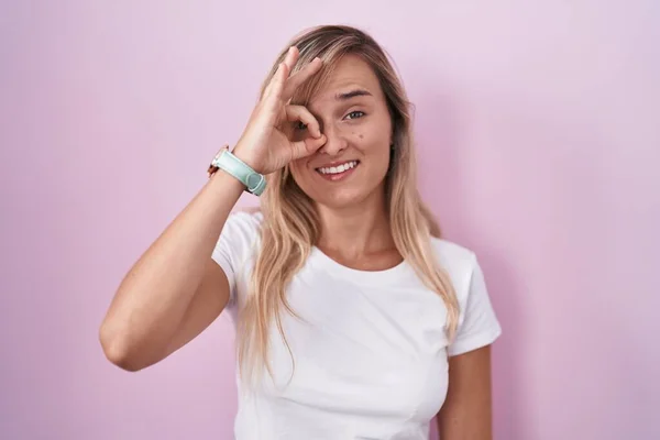 Young blonde woman standing over pink background doing ok gesture with hand smiling, eye looking through fingers with happy face.