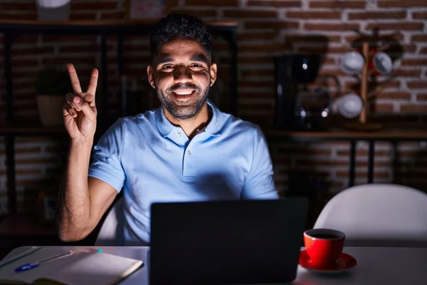 Hispanic man with beard using laptop at night smiling with happy face winking at the camera doing victory sign. number two.