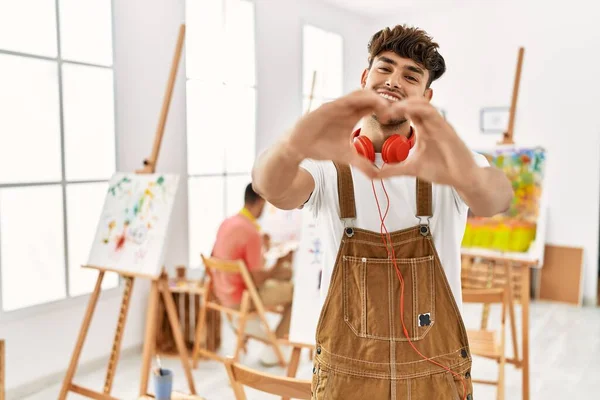 Young hispanic man at art studio smiling in love doing heart symbol shape with hands. romantic concept.