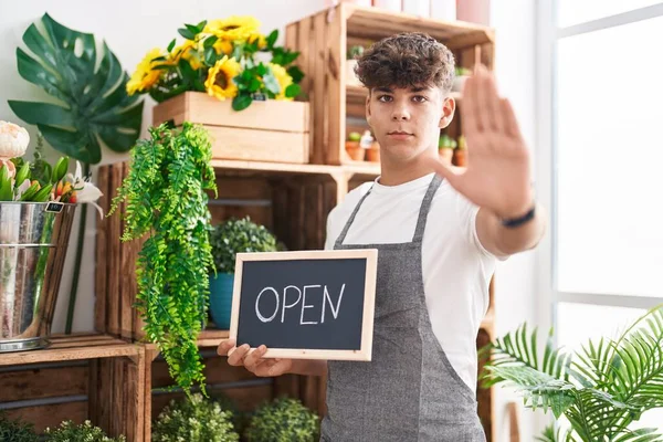 Hispanic teenager working at florist holding open sign with open hand doing stop sign with serious and confident expression, defense gesture