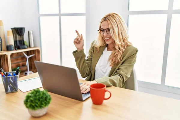 Beautiful blonde woman working at the office with laptop with a big smile on face, pointing with hand and finger to the side looking at the camera.