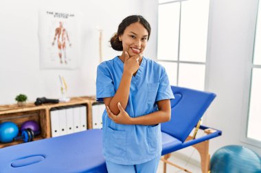 Ağrı kesici kliniğindeki güzel İspanyol fizyoterapist kadın kameraya kollarını kavuşturarak ve çenesini kaldırarak gülümsüyor. Olumlu düşünmek. 